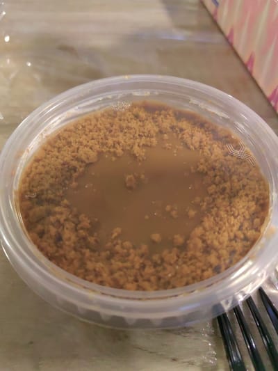 The image displays a close-up, slightly overhead view of a clear, round plastic container holding a two-layered dessert. The bottom layer is a light brown, semi-liquid or viscous substance, partially covered by a top layer of darker brown, coarse, granular crumbs. These crumbs form an irregular perimeter, leaving a central area of the liquid base exposed. Small air bubbles are visible along the inner upper rim of the plastic container. The container itself has a subtle ridged texture on its lower exterior.
The container is situated on a light-colored, possibly wooden, surface. In the upper right background, a portion of a geometric patterned object, likely a box or package, is visible, featuring a design of repeating light pink and white chevron or diamond shapes. To the lower right of the container, segments of a black plastic utensil, likely a fork, with visible tines, are present. A clear plastic film or wrap is also discernable beneath the container, reflecting light and slightly obscuring the underlying surface texture. The scene depicts an indoor environment. The specific location in Kasak, Bulgaria, cannot be determined from the visual information provided within the image.