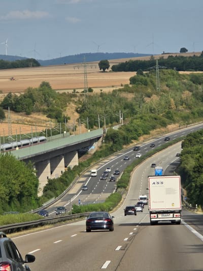 The image depicts a wide, multi-lane highway with several vehicles traveling in both directions. In the foreground, a dark-colored car is visible from the rear, traveling in the right lane. Further ahead, a white semi-trailer truck with a blue trailer is in the left lane, and a white camper van is in the right lane. On the highway to the left, multiple cars are traveling, including a black SUV, a silver sedan, and a white hatchback.
An elevated train track with a modern silver train is visible on a concrete bridge spanning across the highway and the surrounding landscape. The landscape consists of rolling hills covered in green trees and fields of harvested crops, with a few wind turbines visible in the distance on a hilltop. Power lines and electrical towers are also present throughout the scene. The sky is partly cloudy. The image is taken from an elevated perspective, looking down onto the highway and surrounding area. The context of Brechen, Germany, is provided by the presence of a sign with "ASS TRANSPORTE" and a German license plate on the semi-trailer truck, as well as the typical German highway and landscape features.