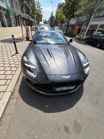 The image shows a dark grey Aston Martin parked on the right side of a street in Erlangen, Germany. The car is facing the camera, and its front is clearly visible. The street has a cobblestone sidewalk to the left, with a black and white bollard on it. In the background, there are buildings with shops and trees lining the street. Other cars, including a black SUV, are also visible on the street. The sky is blue with some clouds. The license plate of the Aston Martin reads "ER TR 7".