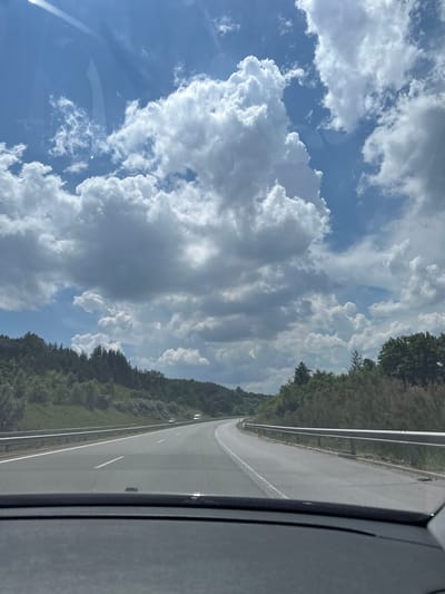 The image depicts a view from inside a vehicle driving on a highway. The road curves to the right and is flanked by a metal guardrail. On either side of the highway are green, tree-covered hills. The sky is blue with large, puffy white clouds, some of which are darker and appear to be gathering. The sun is visible, casting a bright light and creating reflections on the windshield. In the distance, a few vehicles can be seen on the highway. The foreground shows the interior of the car's dashboard. The scene is consistent with a highway drive through a rural, forested area on a partly cloudy day.