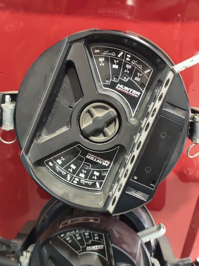 The image shows a close-up, overhead view of a black and red wheel balancing machine with the "HUNTER Engineering Company" logo visible. The machine has several dials and indicators, including icons depicting different vehicle suspension types and tire positions. A metal arm extends from the right side of the machine. The machine is placed on a reflective red surface, likely the floor or a larger piece of equipment. The background is a solid, blurred red. The image was taken in Atameken, Kazakhstan.