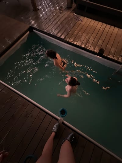 The image captures a nocturnal scene at an outdoor swimming pool, likely in Ivachiv Horishnii, Ukraine, based on the context provided. Two individuals, both appearing to be young women, are in the water. One, further from the viewer, is in a black bikini and is looking away. The other, closer to the viewer, is in a light-colored bikini and is looking upwards. The pool itself is rectangular with a light-colored liner and is filled with clear, dark-green water. The surrounding area consists of a wooden deck, which appears damp, suggesting recent rain or moisture. The deck is illuminated by artificial light, creating reflections on the wet wood. In the foreground, the legs and feet of a person wearing grey Crocs are visible, partially obscuring a blue hose. The overall atmosphere is relaxed and casual, indicative of a private gathering or leisure activity.