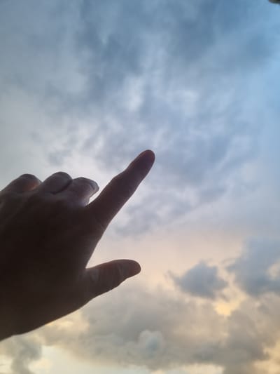The image displays an upward perspective of a cloudy sky, likely at either dawn or dusk, with a human hand prominently featured in silhouette against the bright backdrop. The sky transitions from a soft blue-grey on the left to warmer golden and orange hues on the right, suggesting the presence of a low sun beyond the frame. Clouds of varying densities are scattered across the sky, illuminated from behind to create bright edges and darker, textured bodies.

In the foreground, the hand is positioned in the lower-center, with the index finger extended and pointing towards the upper left portion of the sky. The other fingers are partially visible, curled, and also rendered in deep shadow. The hand's dark silhouette contrasts sharply with the luminous sky, emphasizing the light source behind it. No specific objects or text are visible within the frame apart from the hand and the clouds. The scene conveys an outdoor setting, implying an individual looking up at the vastness of the sky in Kemul, Russia. The act of pointing suggests direction, observation, or perhaps contemplation of the atmospheric conditions.