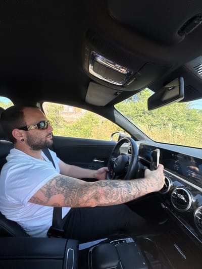 The image shows a man seated in the driver's seat of a car, possibly a Mercedes-Benz based on the steering wheel emblem. He has a beard, is wearing sunglasses, and has extensive tattoos on his left arm. He is holding a smartphone in front of the car's infotainment screen. The environment visible through the windshield suggests an outdoor setting with greenery. It is impossible to independently confirm the location of Nettuno, Italy.