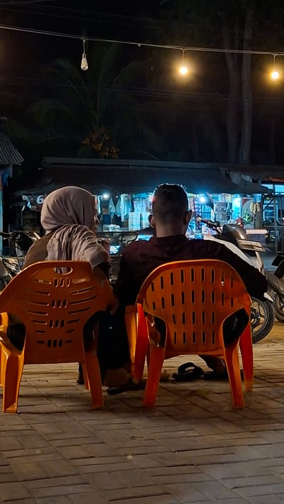 The image depicts two individuals, a man and a woman, seated on orange plastic chairs in an outdoor setting at night. The man is wearing a dark shirt and is looking away from the camera, his profile visible. The woman is wearing a light-colored hijab and is also looking away. They appear to be at a roadside stall or small shop, indicated by shelves with various items behind them. A motorcycle is parked nearby. The ground is paved with rectangular tiles. Two warm-toned lights hang from wires above, illuminating the scene. The overall atmosphere is casual and seems to capture a moment of rest or conversation. The time of day is clearly night. The location is identified as Keudeteupinpunti, Indonesia. There is no visible text in the image.