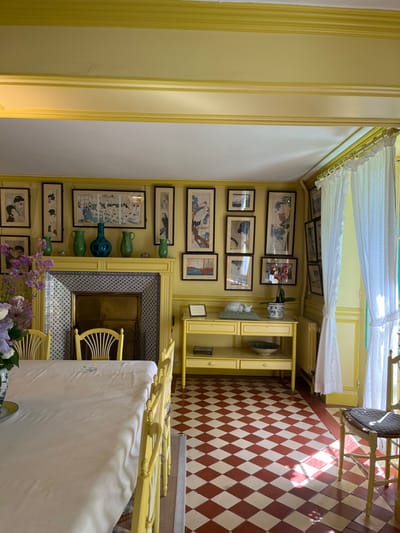 The interior of a room in a house in Giverny, France, located at latitude 49.076 and longitude 1.534. The room is painted yellow, and the walls are adorned with numerous framed prints, primarily of Japanese art depicting birds and nature scenes. A fireplace with a tiled surround sits to the left, and a wooden table with two shelves sits to the right. A white porcelain cat sits on the top shelf of the table. The room has a checkered tile floor in shades of red and cream, and a large white tablecloth sits on a table with four yellow chairs. There is a single white chair with a straw seat on the far right of the room. A window with white lace curtains provides light to the room, and a potted plant with pink flowers sits on the table.  There is a small table with a white vase on top, and the room is otherwise empty.
