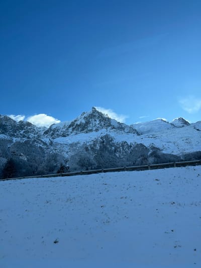 This image captures a magnificent winter mountain landscape under a clear blue sky, set in Les Houches, France.
The scene is dominated by towering, rugged mountains extensively covered in brilliant white snow and ice. These snow-capped peaks stretch across the middle and right side of the frame. Darker, rocky sections and sparse conifer trees, also dusted with snow, are visible on the slopes, providing contrast to the pristine white. Wisps of cloud or wind-blown snow cling to some of the higher ridges, adding to the dynamic feel of the high-altitude environment.
A vast, deep blue sky fills a significant portion of the upper left, indicating a bright, sunny day. The sunlight strongly illuminates the snow, creating a crisp and cold atmosphere. A few thin, white clouds are scattered in the sky, particularly near the mountain tops.
In the foreground, a flat, snow-covered surface occupies the right third of the image. A dark, thin metal railing or barrier, possibly part of a viewing platform, balcony, or mountain road, runs horizontally across this snow-covered ground. The snow on this surface is speckled with small dark specks, likely dirt or debris.
No people or explicit activities are visible, suggesting a serene moment of observation of the natural grandeur. The image evokes feelings of awe, tranquility, and the pristine beauty of an alpine winter. The clear conditions suggest mid-morning or early afternoon. There is no visible text in the image.