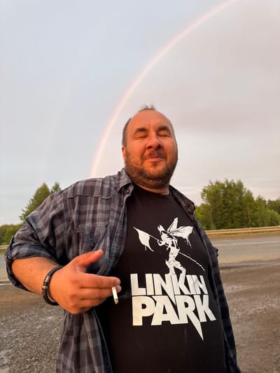 The image depicts a man standing outdoors, holding a lit cigarette in his right hand. He is wearing a dark t-shirt with a white graphic and text that reads "LINKIN PARK." Over the t-shirt, he has a grey and black plaid button-up shirt, unbuttoned. He has a beard and is looking slightly down and to his left with his eyes closed, a faint smile on his face. A rainbow is visible in the sky to the left of the frame. The background shows trees and a road. The sky is overcast, suggesting it might be after rain. The lighting indicates it is likely late afternoon or early evening. The setting appears to be a rural or semi-rural area, consistent with the provided location context of Sovkhoznyi, Russia. The man's relaxed posture and closed eyes suggest a moment of contemplation or enjoyment. He is wearing a dark bracelet on his left wrist.