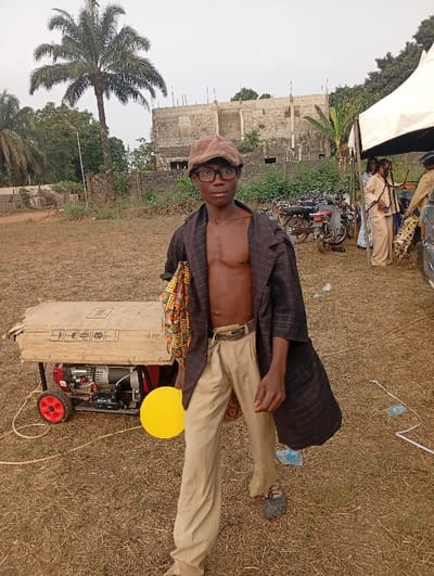 A skinny person wears glasses, a newsboy cap, tan pants, a belt, and a brown blazer. The person's bare chest is visible. They hold a yellow disc. Behind them is a generator with red wheels, motorcycles, a white tent, people, a palm tree, and an unfinished building. The ground is dry grass.
