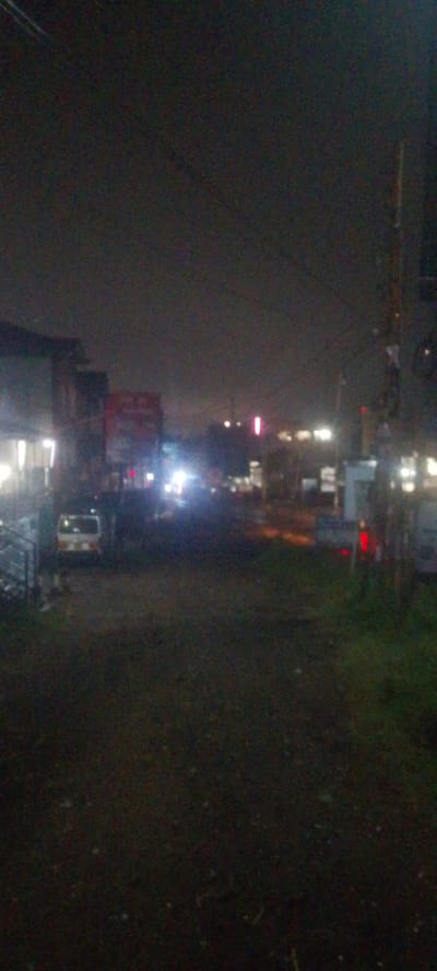 The image depicts a street scene in Ndenderu ward, Kenya, at night. Illumination comes primarily from streetlights and vehicle headlights. Buildings are visible on either side of the street, with a red sign prominent on one structure. Several parked cars line the sides of the road. The road surface appears dark and somewhat uneven. Vegetation, including grass, grows along the edges of the street.