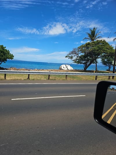 The image depicts a scenic coastal view, likely from a moving vehicle in Lahaina, Hawaii. A bright blue sky with scattered white clouds stretches above a calm, azure ocean. The coastline is lined with green grass and trees, with a prominent shipwreck of a white boat visible on the shore in the mid-ground. A metal guardrail separates the road from the coastal area. The foreground shows a section of asphalt road with a white line marking the lane. A black side mirror of the vehicle is visible in the lower right corner, reflecting a yellow road marking and the ocean beyond. The overall impression is one of a sunny day with a touch of maritime history.