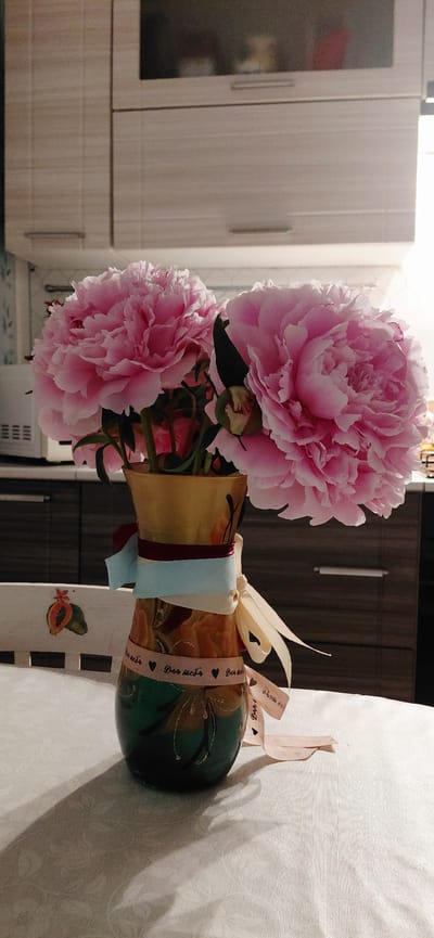 In a domestic indoor setting, likely a kitchen, a vase filled with lush pink peonies is prominently displayed. The vase is a tall, slender, golden-hued vessel with a green base, adorned with decorative painted elements and ribbons. Three ribbons are tied around the neck of the vase: one deep red, one light blue, and one cream-colored. A fourth pink ribbon, inscribed with the Russian phrase "для тебя" (for you) and small hearts, wraps around the lower portion of the vase. The flowers, with their abundant petals, are the clear focal point, casting soft shadows on the white, textured tablecloth. Behind the vase, kitchen cabinets with light-colored wood grain are visible, along with the corner of a white appliance. The overall scene evokes a sense of thoughtful gifting or a celebration. The location is identified as Tashkazgan, Russia.