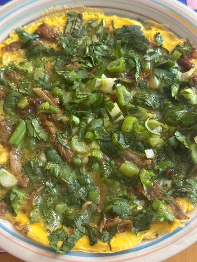 An overhead, close-up shot captures a savory dish served on a light-colored plate. The dish features a round, golden-yellow base, possibly an omelette or a thin, egg-based pancake. It is generously covered with a vibrant mix of fresh green ingredients, predominantly chopped green onions (scallions) and cilantro leaves, providing varying shades of green and texture. Beneath and intertwined with the green toppings, darker, brownish, shredded elements are visible, which could be pulled meat, mushrooms, or another savory filling. The surface of the dish appears moist and glossy, suggesting the presence of a sauce or liquid. The food is presented on a ceramic plate with a subtle, striped rim displaying faint lines in colors such as pink, blue, and beige. No activity or people are depicted in the image. There is no visible text. The time of day, weather, and specific location cannot be determined from the visual content.