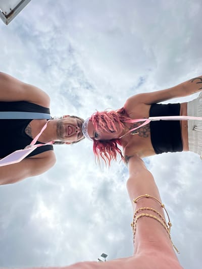 A low-angle shot depicts two people in Kampung Toyapakeh, Indonesia. One person is upside down, wearing a white top, black shorts, and has pink hair. They have tattoos on their arms and chest, and are wearing a belly button piercing. The other person is right-side up, wearing a black tank top, and sticking their tongue out. Both are wearing pink lanyards. A hand wearing gold bracelets is visible on the right side of the frame. The background features a cloudy sky.