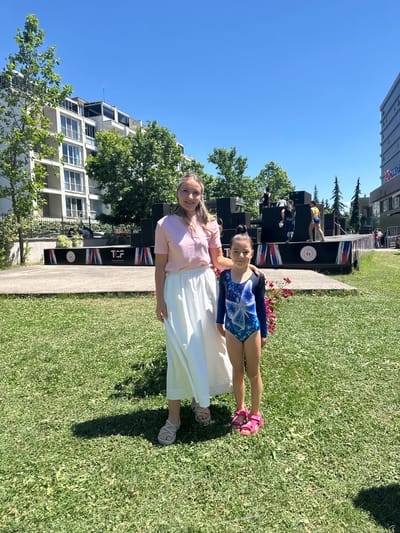 A woman and a young girl are standing on a grassy area in front of a stage. The woman is wearing a pink t-shirt and a long white skirt, and the girl is wearing a blue and white gymnastics leotard. The stage is decorated with flags and logos, and there are several people on it, some of whom appear to be performing. In the background, there are buildings and trees, indicating an urban environment. The scene appears to be a public event, possibly a gymnastics competition or performance, taking place outdoors on a sunny day. The location is the Sancaktepe City Square in Istanbul, Turkey.
