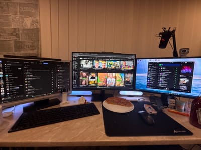 The image depicts a desk setup with multiple computer monitors, a keyboard, a mouse, and a microphone. The monitors display various applications, including what appears to be Discord, YouTube, and Spotify. On the desk, there is a slice of pizza on a plate, a bottle of ketchup, and some small figurines. The background shows a wall with a textured pattern and blinds. The lighting suggests it is either daytime with ambient light or nighttime with artificial lighting. The scene is indoors, likely in a home office or gaming setup. No specific location in the city of Beloslav, Bulgaria can be identified from the visual elements of the image.