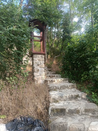 The image shows a stone staircase leading upwards through dense vegetation. At the base of the stairs, to the left, are black trash bags and dry grass. The stairs themselves are made of unevenly cut stone slabs. To the left of the stairs, a stone pillar supports a wooden structure with an arched opening. Behind the pillar and to the left of the stairs, there are remnants of a stone wall. The environment is lush and overgrown with trees and bushes. The lighting suggests it is daytime, likely late afternoon due to the shadows. The scene is set in an outdoor, natural environment. This scene is located in Melnik, Bulgaria, specifically at the Stairs to the Kordopulov House.