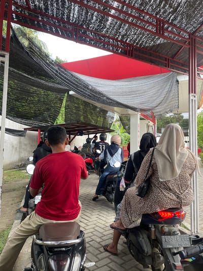 The image depicts a line of motorcycles with riders waiting under a covered structure, likely a gas station canopy, in Glee Gogo, Indonesia. A man in a red shirt is in the foreground, sitting on a black Honda scooter. Behind him, several other individuals are on their motorcycles, some wearing helmets. The structure's roof is made of dark netting stretched over a red metal frame. To the left, a concrete wall is visible, and to the right, part of a building with red accents is present. The ground is paved with interlocking bricks. The overall impression is of a typical scene at a local service station during a potentially busy period.