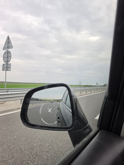 View from inside a car traveling on a highway in Vinarovo, Bulgaria. A side mirror reflects the road behind, showing lane markings and other vehicles. An arrow-shaped road sign is visible in the distance along the roadside. The sky is overcast.