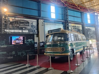 The image shows an interior view of a museum or exhibition space, likely the Museum of the Macedonian Railway or a similar transportation-themed exhibit in Menemeni Municipal Unit, Greece. In the foreground, a vintage green and tan bus, possibly a Mercedes-Benz O 302, is parked on a red carpeted area, cordoned off by silver stanchions with black velvet ropes. The bus has a sign on its front that reads "ΘΕΣΣΑΛΟΝΙΚΗ". Behind the bus, large black display panels are mounted on the wall, featuring historical photographs of buses and timelines with dates such as 1900, 1929, 1930, 1940, and 1953. One panel above the bus shows a black and white image of an older bus with the Greek text "ΤΑΞΙΔΕΥΟΥΜΕ ΜΑΖΙ" (We travel together). Another display behind it shows a large black and white photo of a bus with the text "ΧΟΡΗΓΟΣ: ΣΤΑΘΜΟΣ ΜΑΚΕΔΟΝΙΑ ΥΠΕΡΑΣΤΙΚΩΝ ΛΕΩΦΟΡΕΙΩΝ ΚΤΕΛ" (Sponsor: Macedonia Intercity Bus Station KTEL). The space has a high industrial ceiling with exposed metal beams and blue corrugated metal walls with large windows. A single person, wearing a white t-shirt and shorts, is visible walking away from the camera on the right side of the frame.
