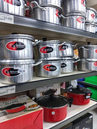 The image displays a variety of cookware on shelves. The top two shelves hold stacks of aluminum pots with handles and lids. Each pot features a red and black "18 Jawa CM Mashion Group" label. Price tags reading "PANCI JAWA J" and "37.000" are visible. The bottom shelf showcases red and black Dutch ovens and soup pots, along with boxes labeled "OVEN" and "SOUP POT." Green plastic trays are also present on the shelves.