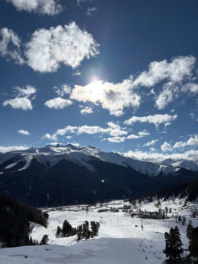 The image displays a wide panoramic view of a snow-covered mountain landscape and ski resort located in Lunnaya Polyana, Russia. The scene is illuminated by bright sunlight under a clear, deep blue sky dotted with scattered white cumulus clouds. The sun, partially obscured by a cloud in the upper-center, produces a noticeable flare with iridescent spectral effects.
In the mid-ground, a prominent range of rugged mountains is visible, with their upper sections covered in white snow and lower slopes densely forested with dark evergreen trees. Some clearings and potential ski trails are discernible on these higher slopes.
The foreground and lower mid-ground reveal a broad valley floor entirely blanketed in snow, indicative of a winter resort. Multiple groomed ski slopes with discernible tracks are present. Several ski lift systems, including poles and lines with yellow chairs, traverse the slopes. A cluster of resort buildings, characterized by their low-rise architecture, is situated on the valley floor, primarily on the right side. Scattered groups of evergreen trees, some appearing tall and dark against the snow, punctuate the landscape. Small, indistinct figures, consistent with skiers or snowboarders, are visible on various slopes throughout the resort area. No specific actions or interactions are clearly identifiable due to the distance. The overall environment suggests a vibrant winter sports destination.