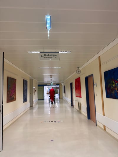 The image shows a well-lit hospital corridor in Amstetten, Austria. A person in a red outfit is being pushed in a wheelchair down the center of the hallway. The walls are lined with artwork. Signage for "Radiologie Aufnahme 2/3" is visible. The floor is glossy, and there are floor markers on the right side of the hall. A clock hangs on the wall near the ceiling.