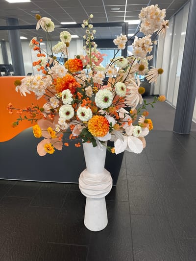 A tall, white, textured vase holding a large arrangement of artificial flowers stands on a dark grey tiled floor in what appears to be an office environment. The flowers are a mix of colors, predominantly white, orange, and pale pink, with some green foliage and small red berries. The arrangement is lush and multi-layered, with some flowers featuring prominent yellow centers. In the background, parts of an orange and grey wall or partition are visible, along with office furniture and glass partitions. The overall lighting suggests an indoor, well-lit space. The specific location in Staphorst, Netherlands is not ascertainable from the visual information provided.
