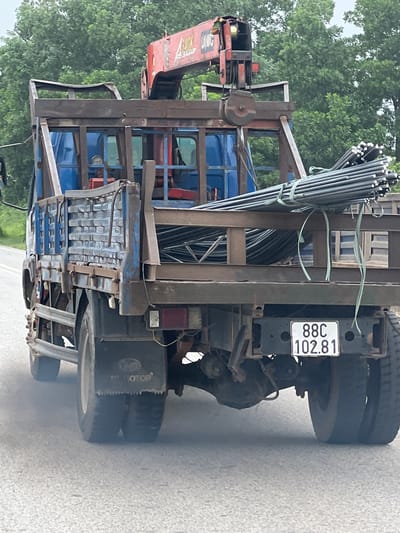 A blue truck with a red crane attachment is shown from the rear, driving on a paved road. The truck is carrying a load of metal rebar strapped down in the bed. The license plate of the truck reads "88C 102.81". The truck is moving, as indicated by the smoke coming from the rear wheels. Trees are visible in the background, suggesting a rural or semi-rural setting, possibly on the outskirts of Bá Hiến, Vietnam. The sky is overcast. The scene is captured in daylight.