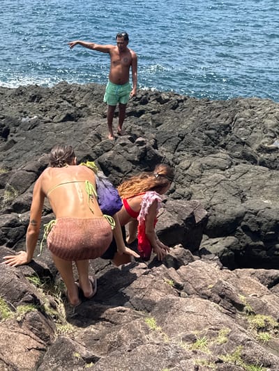 The image depicts a coastal scene with three individuals on a rocky cliff overlooking the ocean, likely in Santa Monica, Philippines. The man on the left stands near the edge, wearing light green shorts, his arm outstretched towards the ocean. Two women are on the rocks. One is wearing a red top and a pink and red garment. The second woman is wearing a bikini with a brown knit skirt. The cliff is composed of dark, jagged rocks, with sparse vegetation growing on the upper portions. The ocean is visible to the left, showing a textured surface with shimmering light.