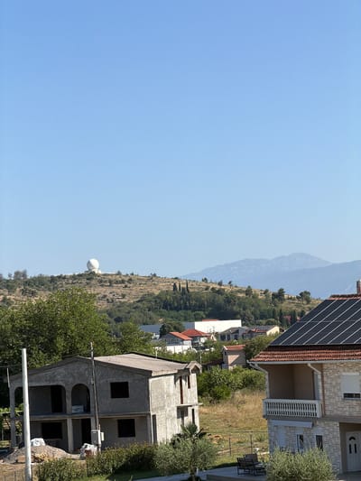 The image displays a rural residential area with a clear blue sky overhead. In the background, a prominent white spherical structure, likely a radar dome, is situated on a rounded hill. The landscape between the foreground and the hill is dotted with scattered trees and dry grass. In the middle ground, several houses of various sizes and architectural styles are visible. One house in the foreground is under construction, with exposed concrete and unfinished features. Another adjacent house is a completed multi-story dwelling with a tiled roof and solar panels. The scene appears to be set in Mojanovići, Montenegro, a location known for its proximity to Podgorica and its developing residential areas. The overall impression is one of a sunny day in a developing community.