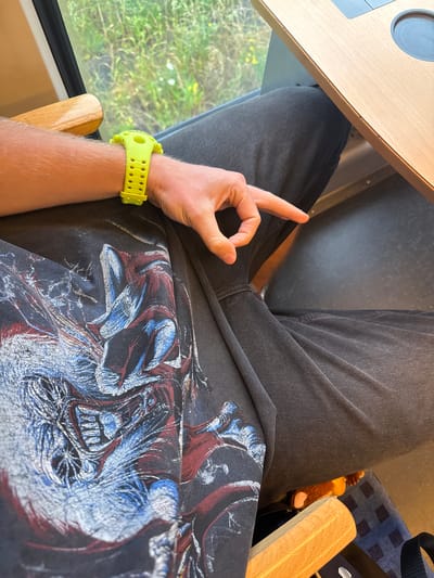 The image shows a person wearing a black graphic t-shirt and dark grey jeans. The person has a neon yellow watch on their left wrist and is making the "circle game" gesture with their right hand, with the thumb and index finger forming a circle. The person is sitting in a train or bus, with a wooden table in front of them and a window to their left showing green foliage and some yellow flowers. The background is blurred. The image does not contain any information about the city of Bitterfeld-Wolfen, Germany.