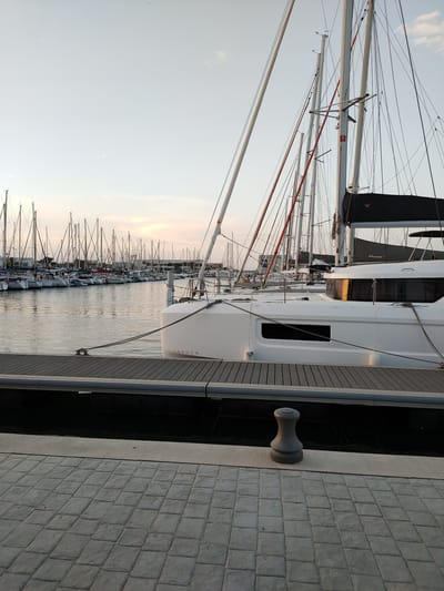 The image depicts a marina in Canet-en-Roussillon, France, likely during the late afternoon or early evening, given the soft, diffused light. The foreground is dominated by a paved walkway with a patterned, rectangular stone surface. Adjacent to the walkway is a wooden dock, also with a patterned surface, extending into the water. A dark grey bollard is positioned on the edge of the dock.

In the water, several sailboats are moored. The closest and most prominent is a white catamaran with the word "LAGOON" visible on its hull. This boat is tied to the dock with thick ropes. Behind this vessel, a multitude of other sailboats, mostly white, are lined up, their masts creating a dense forest of vertical lines against the sky. The water in the marina is calm, reflecting the sky and the boats.

The sky is a pale, hazy blue, with a faint suggestion of clouds or atmospheric haze, particularly towards the horizon where a pale sunset glow is visible. The overall impression is one of tranquility and order, characteristic of a well-maintained marina. There are no people visible in the image.