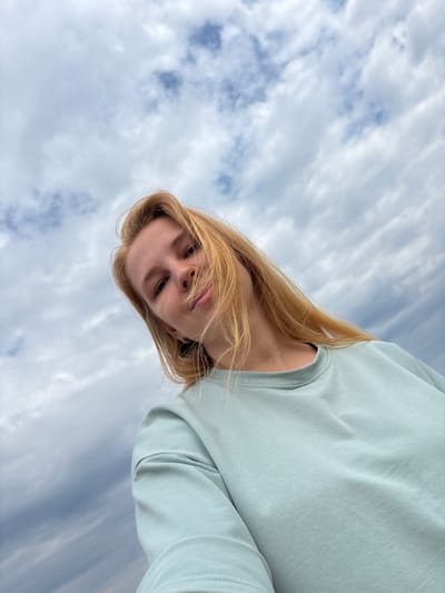 A low-angle, close-up selfie of a young Caucasian woman with shoulder-length blonde hair against a cloudy sky. She is wearing a light blue crew-neck sweatshirt. The image appears to be taken outdoors, possibly in the city of Breytovo, Russia, given the information provided. The sky is filled with dense, gray and white clouds, suggesting an overcast day. Her hair is slightly windswept, and she is looking towards the camera with a neutral expression.