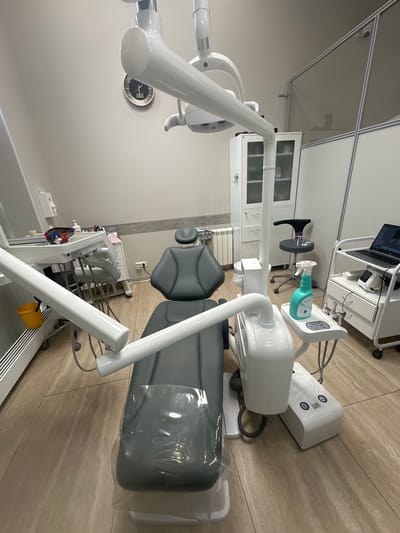 The image shows a dental examination room. The room is clean and modern. A dental chair is the main subject, covered in clear plastic. Behind it is a wall-mounted instrument and a cabinet of white drawers. To the left is the dentist's lamp and clock mounted to the wall. To the right, sits a small cart carrying a laptop, and a storage cart of dental tools sits behind the chair. The floor is covered in beige tiles. The location provided is Kraskovo, Russia. There are no people present.