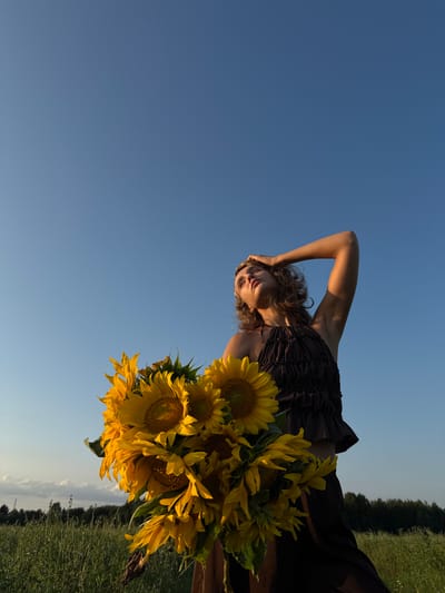In Aksaitovo, Russia, a young woman stands in a field of grass, holding a large bouquet of sunflowers. She is wearing a dark brown, ruffled halter top and a dark skirt. Her hair is curly and blonde, and she is looking up at the sky with her right hand on her forehead. The sky is a clear, bright blue, and the sun is shining. In the background, there is a line of trees. The overall impression is one of a serene and beautiful summer day.