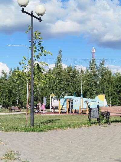 The image depicts a bright, sunny day in a park, likely in Roshchinsky, Russia. The scene features a playground with a blue and yellow airplane-shaped structure, complete with slides and climbing elements. Several people, including children and adults, are present on or near the playground. One child in a pink shirt and shorts stands near the airplane structure, while others are visible in the background.
In the foreground, a paved walkway leads into the park. A lamppost with two spherical lights stands prominently on the left side of the image, with a young tree growing beside it. To the right, a wooden bench is visible, partially obscured by a black trash can. Further in the background, a dense line of tall, green trees forms a backdrop to the park. A cellular tower is visible in the distance, rising above the trees. The sky is a vibrant blue, dotted with fluffy white clouds, suggesting fair weather. The overall atmosphere appears relaxed and cheerful, indicative of a typical summer day spent outdoors.