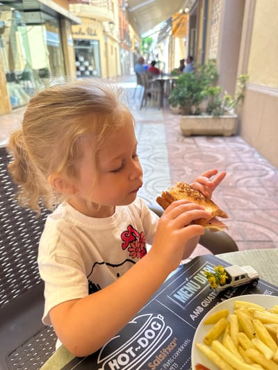 A young Caucasian girl with blonde hair pulled into a ponytail is seated at an outdoor cafe table. She is wearing a white t-shirt with a floral design. In her hands, she holds a half-eaten sandwich, which appears to be a grilled cheese or ham and cheese sandwich. In front of her on the table is a menu, a small toy white pickup truck adorned with yellow wildflowers, and a plate of french fries. The background shows a narrow street lined with buildings, suggesting an urban or town setting. The architecture and awnings are typical of Mediterranean coastal towns. The specific location is Sant Feliu De Guíxols, Spain, a coastal town in Catalonia. The girl's attention is focused on the sandwich as she holds it.