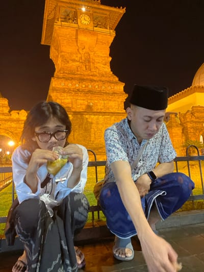 Two individuals are squatting at night in Kauman, Indonesia, against an illuminated architectural backdrop. On the left, a person with long dark hair and glasses wears a white long-sleeved shirt, a dark patterned sarong, and dark flip-flops, actively consuming a yellowish substance from a clear plastic pouch. A lanyard with a white ID card is visible. On the right, an adult male wearing a black peci, a light-colored patterned short-sleeved shirt, a blue patterned sarong, and grey slides is observed looking downwards, with his right arm extended towards the ground. A dark smartwatch is on his left wrist, and a crossbody bag strap is visible. Behind them, a dark metal fence separates the foreground from the brightly lit brick-colored clock tower of the Great Mosque of Kauman, which displays a clock face indicating approximately 10:55. To the right of the tower, part of a white domed structure is also illuminated.