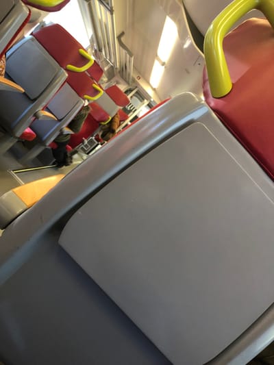 The image displays the interior of a passenger train carriage, observed from a tilted perspective, ostensibly within the city of Dettwiller, France. The environment is characterized by rows of train seating. The seats are primarily red with light gray plastic backings and armrests. Several yellow, curved grab handles are affixed to the top of the seatbacks throughout the visible section of the carriage. One seat in the left foreground exhibits a yellow-orange accent on its lower back. Overhead, a metallic luggage rack system runs along the ceiling, alongside rectangular lighting fixtures illuminating the compartment. Partially visible are several occupants. On the left side, an individual is seated, dressed in dark clothing, with a backpack visible at their feet. Further down the aisle on the right, another individual is seated, partially obscured, appearing to wear brown, fur-textured outer clothing. No direct interactions or specific actions beyond sitting are discernible. The floor surface is light-colored. Minor scuff marks are present on some of the light gray plastic seat surfaces.