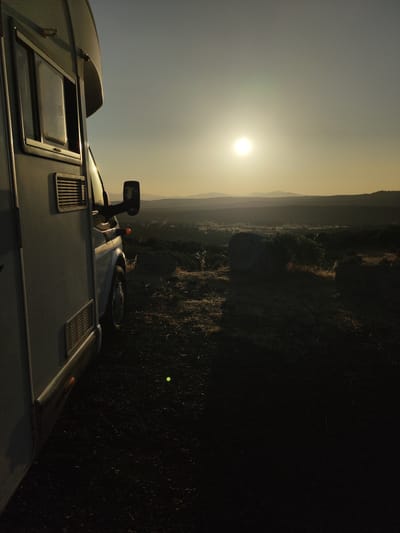 An exterior, low-angle, right-facing shot captures a side view of a light-colored camper van parked on a dirt and rocky surface at dusk. The sun, a bright orb in a hazy sky, is positioned high and slightly to the left of the frame, casting a warm, golden light that illuminates the scene. The camper van occupies the left portion of the image, its window and exterior vents visible. The right side of the image opens to a vast landscape, characterized by rolling hills and mountains in the distance, receding into soft focus. The foreground consists of rough terrain with scattered rocks and dry vegetation, some of which are illuminated by the setting sun. A large, rounded boulder is prominent in the middle ground to the right, partially obscured by a bushy plant. The overall atmosphere is serene and contemplative, suggesting a moment of peaceful repose in a remote natural setting. This scene is located in the municipality of Ledrada, Salamanca, Spain.