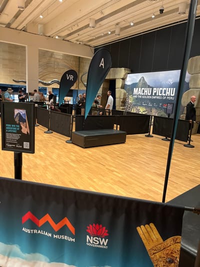 An indoor scene depicts an exhibition space, likely within the Australian Museum in Woolloomooloo, Australia. The area features a polished wooden floor and high ceilings with architectural details and lighting.

The central focus is a large banner advertising "MACHU PICCHU AND THE GOLDEN EMPIRES OF PERU," displaying an image of the iconic Peruvian archaeological site. Adjacent to this, partially visible, are faux stone structures resembling ancient ruins, enhancing the exhibition's theme.

To the right, a tall banner features the "AUSTRALIAN MUSEUM" logo, stylized as a red 'M' or mountain range, and the "NSW GOVERNMENT" logo. This banner also shows a golden, gloved hand artifact against a blue background with clouds.

The space is organized with black stanchions and barriers, suggesting a queue or a defined pathway for visitors. Two prominent tear-drop shaped signs are visible: one labeled "VR" for Virtual Reality and another marked "A," possibly for Augmented Reality or another interactive section.

Further into the foreground, a digital sign on a stand displays "PERU: WIN THE ADVENTURE OF A LIFETIME!" accompanied by a QR code, indicating a promotional competition. A black bench-like object is situated behind the barriers.

Several people are scattered throughout the area. A man in a dark suit stands near the Machu Picchu banner, possibly staff or security. Another man in a white shirt and dark pants is visible near the "A" sign. Other visitors are seen seated or standing in the background, appearing engaged with the exhibition or waiting for access to interactive experiences. The overall atmosphere suggests an organized and engaging museum event.