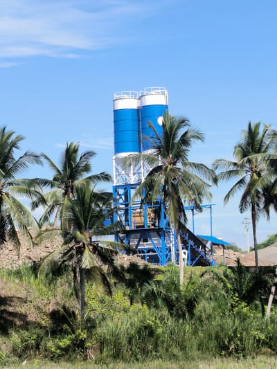 This media file presents an industrial cement batching plant set against a clear blue sky, partially framed by lush tropical vegetation.
The central subject is the multi-component cement batching plant, featuring two prominent, tall cylindrical silos painted in bright blue with white tops and railings. These silos are supported by an intricate blue and white metal framework. Below the silos, various mixing units and conveyor belt structures are visible, also in coordinating blue and white. A circular white emblem or logo is discernible on the side of the left silo, though any specific text on it is unreadable.
The plant is partially obscured by numerous tall palm trees with vibrant green fronds, which occupy the foreground and midground. Dense green undergrowth and tall grasses are visible at the base of the image. To the right of the plant, a large pile of what appears to be construction aggregate (likely gravel or sand) and a utility pole are present. A stone retaining wall or embankment can also be seen behind some of the foliage.
The scene is captured outdoors during the daytime under bright, clear conditions, indicated by the strong sunlight and the expansive blue sky with only a few wispy clouds. There are no people visible, and no specific activity or event is taking place at the moment of the photograph, suggesting the plant is idle. The presence of numerous palm trees strongly suggests a tropical environment, consistent with the specified location of Seuneubok, Indonesia. The image objectively depicts an industrial facility amidst a natural, green landscape.