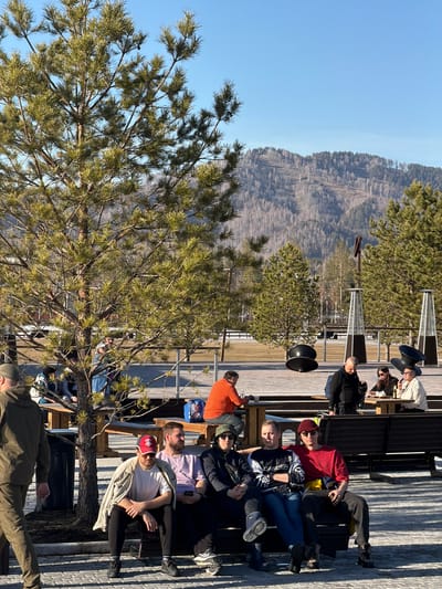 A group of approximately 14 people sit on benches on a wooden deck, overlooking a mountainous terrain under a clear blue sky. The scene is situated in Озёрное, Russia. The people are casually dressed and appear to be relaxing and engaging in conversation. Several industrial-looking speakers and heating units are visible in the background, along with pine trees.