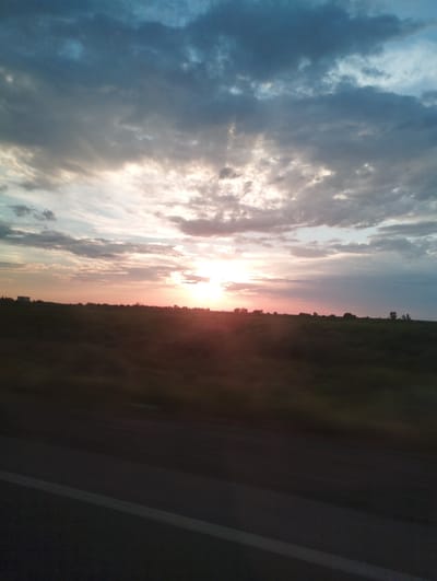 The image depicts a natural landscape scene, possibly taken from a moving vehicle. A sunset or sunrise dominates the sky, with warm hues of orange and pink mingling with grey and blue clouds. Rays of light break through the clouds, illuminating the horizon. The foreground consists of a dark, blurred expanse, suggesting a road surface with a white line marking, and beyond that, a dimly lit field or meadow. A line of silhouetted trees and structures is visible against the sky on the horizon. The overall impression is one of a serene, transitional moment in the day. The location, Ahunbabaev, Uzbekistan, is not visually evident in the image itself.