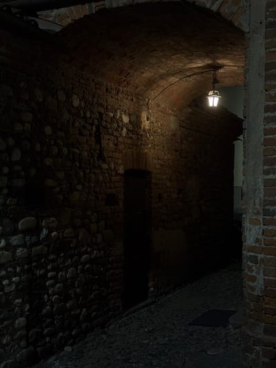 The image shows a narrow, cobblestone alleyway under a brick archway in Lagnasco, Italy. A single, illuminated lantern hangs from the ceiling of the archway, casting a warm glow on the textured brick and stone walls. The alley is dimly lit, with shadows obscuring much of the background. To the right, a portion of a brick building with a weathered concrete pillar is visible. The ground is paved with irregular cobblestones, and a dark rectangular shape, possibly a drain cover, is visible in the middle of the alley. The overall impression is one of a historic and atmospheric passage.