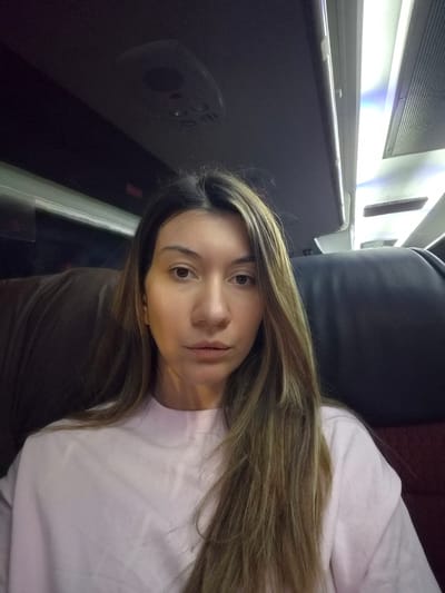 The image displays a female individual, positioned from the chest up, centered in the frame. She has long, straight brown hair with lighter highlights and wears a light pink high-necked, long-sleeved top. Her gaze is directed straight ahead, with a neutral expression. She is seated within the interior of a passenger transport vehicle, likely a bus or train, located in the Municipal Unit of Lamia, Greece. Dark, upholstered seating is visible on both sides of the subject; a brown seat back is to her immediate left, and a dark grey/black seat back is to her right. The overhead ceiling is dark, featuring integrated artificial lighting, including a bright linear light strip along a panel edge and a recessed rectangular light fixture with a grated cover, typical of public transportation. The scene is illuminated solely by artificial light sources originating from within the vehicle. No other individuals or specific actions are depicted.