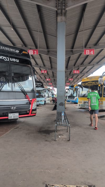 The image depicts a bus terminal in Phakhao, Laos, with several buses parked under a covered structure. The terminal appears to be outdoors, with a concrete floor and a corrugated metal roof supported by metal beams and a central pillar. Red signs with white lettering, indicating bus bay numbers such as "B3," "B4," "B5," "B6," "B7," "B9," "B10," "B11," and "B12," are suspended from the roof.

In the foreground, a black and red bus is parked on the left, displaying a license plate with the characters "UN 3033." To the right, a yellow bus with "VIP Limousine" written on it is visible. A person wearing a green shirt with the number "11" and dark shorts, along with red flip-flops, is walking away from the camera towards the yellow bus. A metal bench with a water bottle on it is positioned between the foreground bus and the yellow bus. In the background, other buses, including a light blue one, are parked, and several people are seen near the buses, some appearing to be passengers or staff. The overall environment suggests a functional transportation hub.