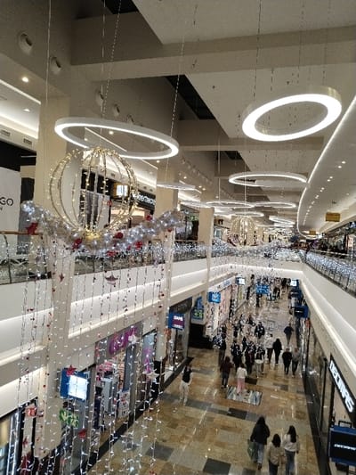 A brightly lit, modern shopping mall is captured from a high angle, revealing a bustling scene. People stroll through the open corridors lined with retail stores. The mall is adorned with festive decorations, including illuminated spheres and cascading string lights. Circular light fixtures hang from the high ceiling, adding to the mall's contemporary ambiance. The floor is tiled with a pattern of dark and light squares.
