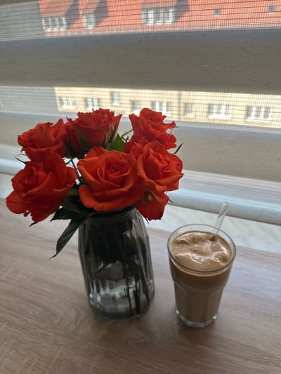 The image captures an indoor scene in the city of Arzberg, Germany, looking out a window onto an urban environment during daytime. On a light-colored wooden surface, likely a table or windowsill, a clear fluted glass vase filled with water holds a bouquet of seven or more vibrant red-orange roses, displaying green stems and leaves. To the right of the vase, a clear glass contains a light brown, frothy beverage, likely an iced coffee or similar drink, with a clear, straight straw inserted into it. The background features a window with a partially drawn, light-colored fabric blind, which filters and blurs the view beyond. Through the translucent blind, reflections or faint outlines of multi-story buildings are discernible, characterized by distinct red-tiled roofs and lighter-toned facades with multiple rectangular windows, suggesting typical residential or commercial architecture. No people are visible, and no actions or interactions are depicted.